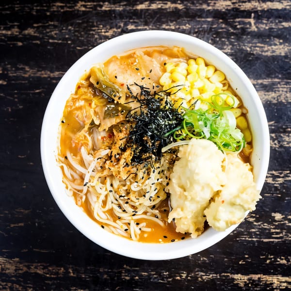 Oki Shōten's ramen bowl with noodles, corn, kimchi, green onions, and tempura on a dark wooden table.